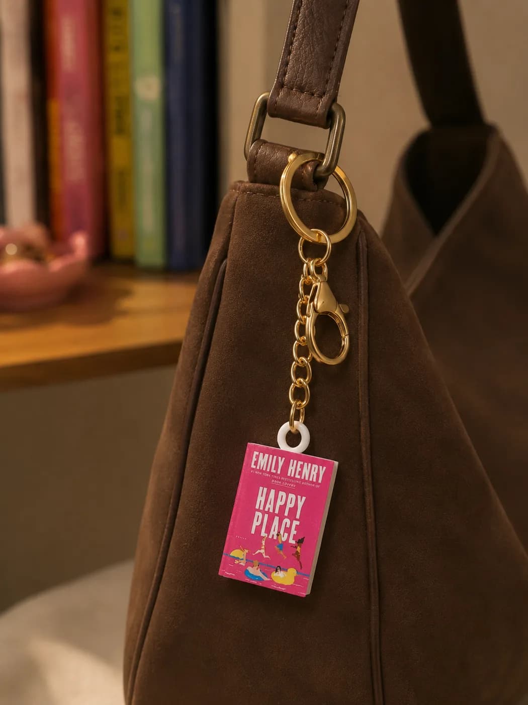 A mini book keychain hanging from the gold strap ring of a brown suede bag, a shelf of books blurred behind