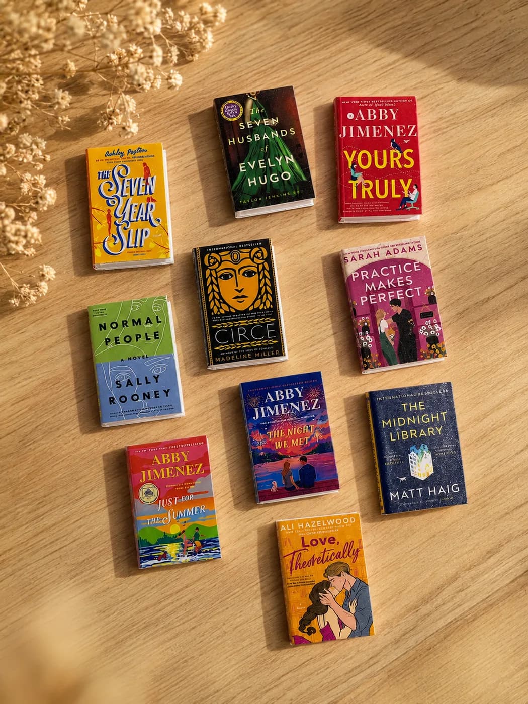 Ten miniature novels spread across a pale wood surface in afternoon light